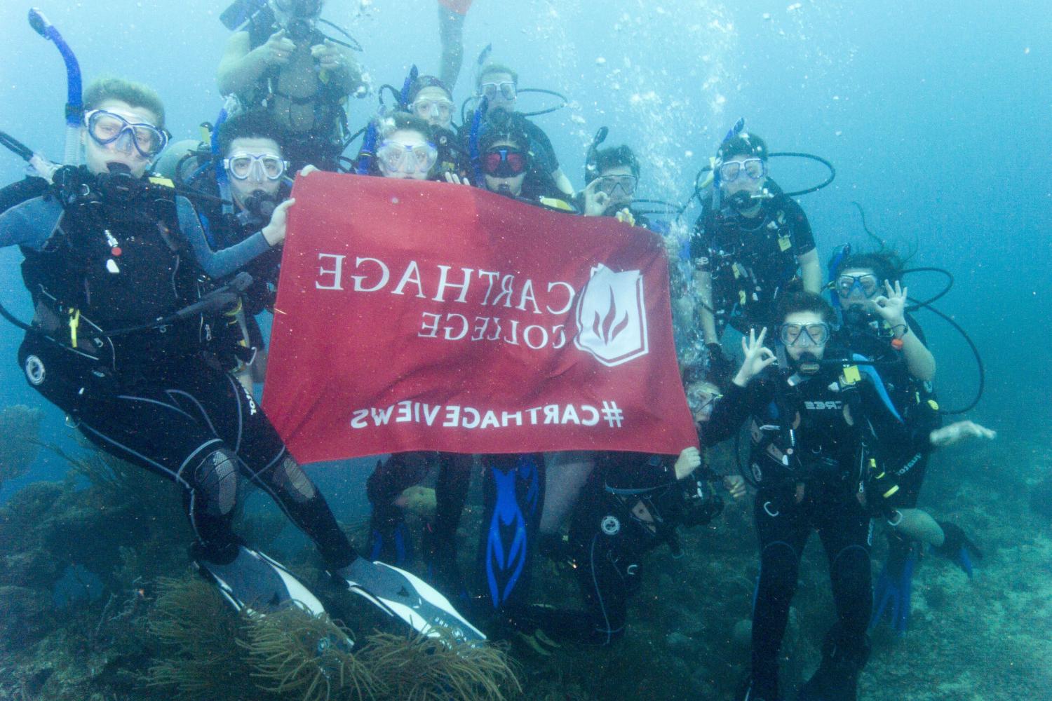 学生们手持<a href='http://2n.awamiwebsite.com/'>bv伟德ios下载</a>旗帜，在j学期洪都拉斯游学之旅中潜水.